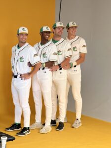 Four baseball players in white uniforms and green hats pose for a photo against a yellow and gray backdrop.