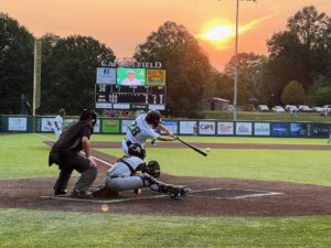 Baseball game at sunset, batter swings, umpire and catcher in position on field.
