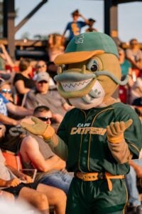 Cape Catfish mascot in green baseball uniform with orange trim, interacting with fans.