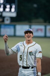 Excited baseball player in white uniform with orange and green trim celebrates with fist pump.