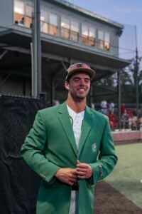 A smiling man in a green Masters jacket and baseball cap stands on a baseball field.