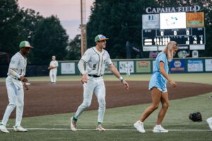 Baseball players walk on field near scoreboard at Capaha Field during a game at dusk.