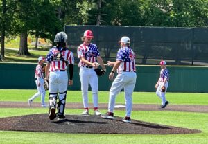 Baseball players in patriotic uniforms on a sunny field, with a pitcher, catcher, and coach interacting.