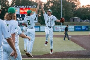 Two baseball players in white uniforms high-five on the field with a scoreboard in the background.