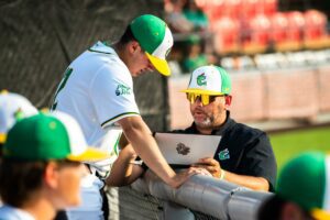 Baseball players in green and white uniforms review a laptop on a sunny day.