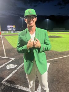 Young man in green jacket and baseball cap holds a baseball on a field at night.