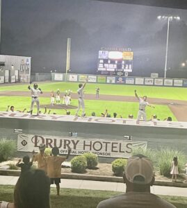 Baseball players celebrate on the field at night in front of a scoreboard and sponsor banners.