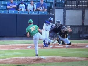 Baseball pitcher in green throws ball to batter with catcher in gear behind home plate.