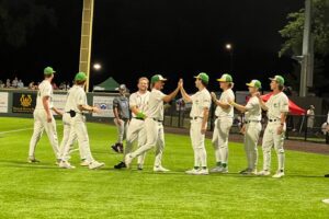 Baseball team celebrates with high-fives on the field at night, wearing green and white uniforms.