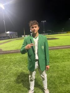 Teen baseball player in green blazer and white uniform on field at night.