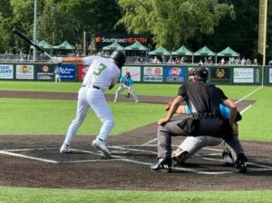 Baseball batter swings at pitch with catcher and umpire behind home plate on a sunny day.