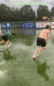 Two boys splash through a flooded baseball field, one in green shirt, the other shirtless, under a cloudy sky.