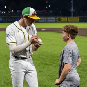 Baseball player signing a baseball for a young fan on the field at night.