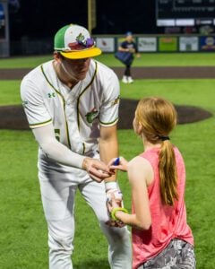 Baseball player signing a baseball for a young fan on a green field at a stadium.