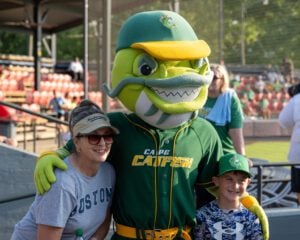 Mascot poses with woman and boy at baseball game, wearing team jersey and hat.