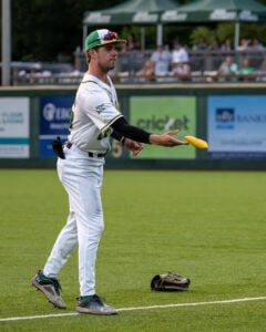 Baseball player in white uniform and green hat tossing a yellow beanbag on a green field.
