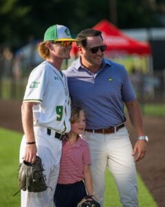 Baseball player and man in Masters polo smile with a child between them on a field.