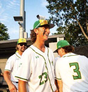 Baseball players in green and yellow uniforms smile and laugh outdoors with a security camera in background.