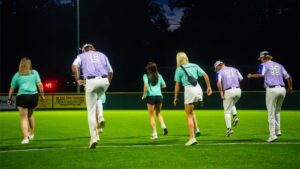 People in sports uniforms run on a bright green field at night, with dark trees in the background.