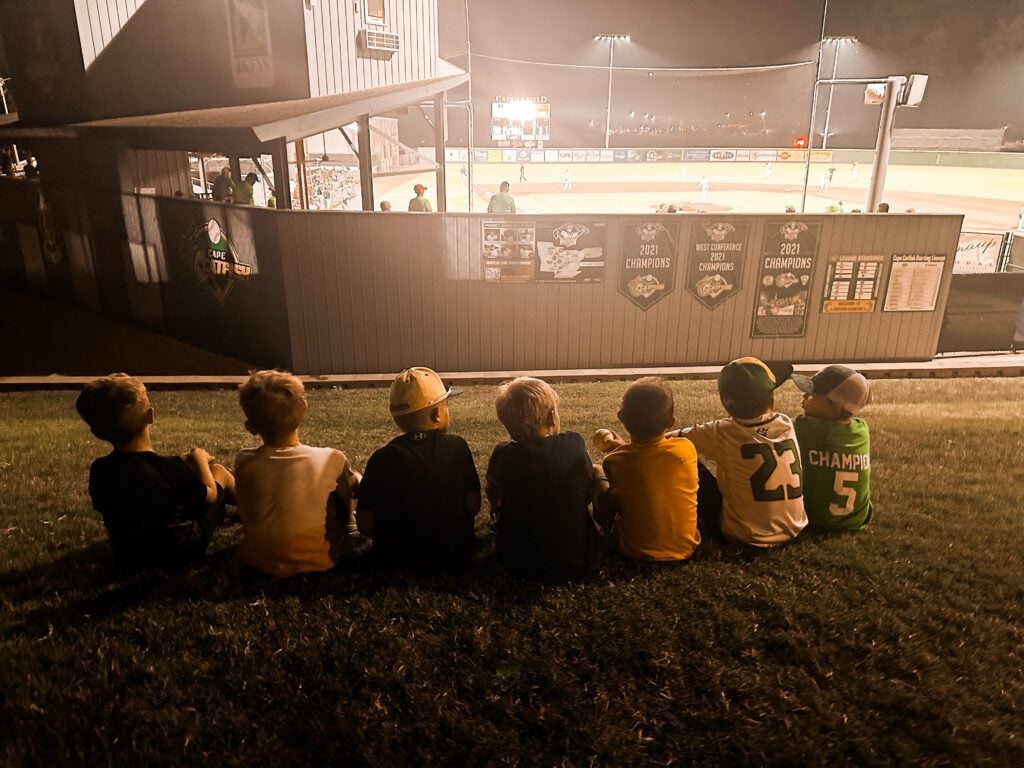 Boys in baseball jerseys watch a game from the grass at night.