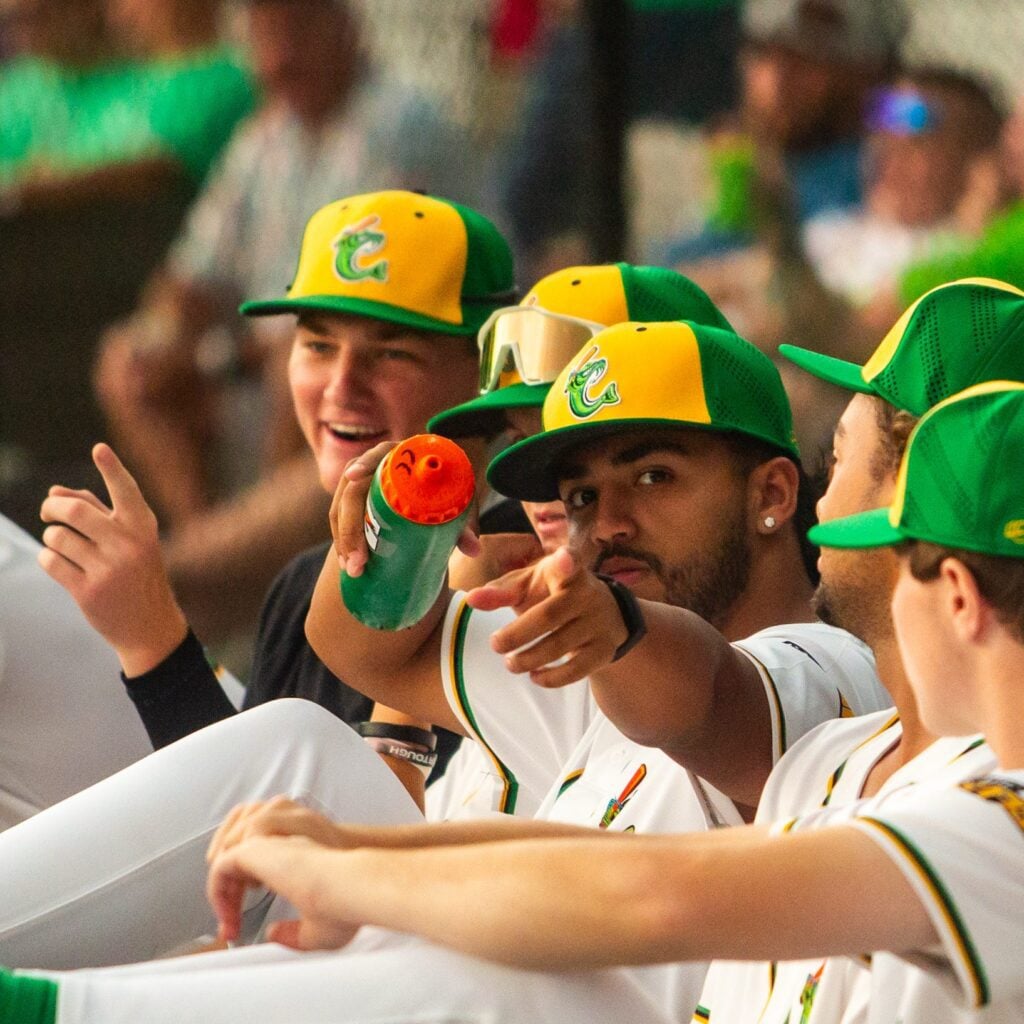 Baseball players in yellow and green hats sit on a bench, one points at the camera, another holds a drink.