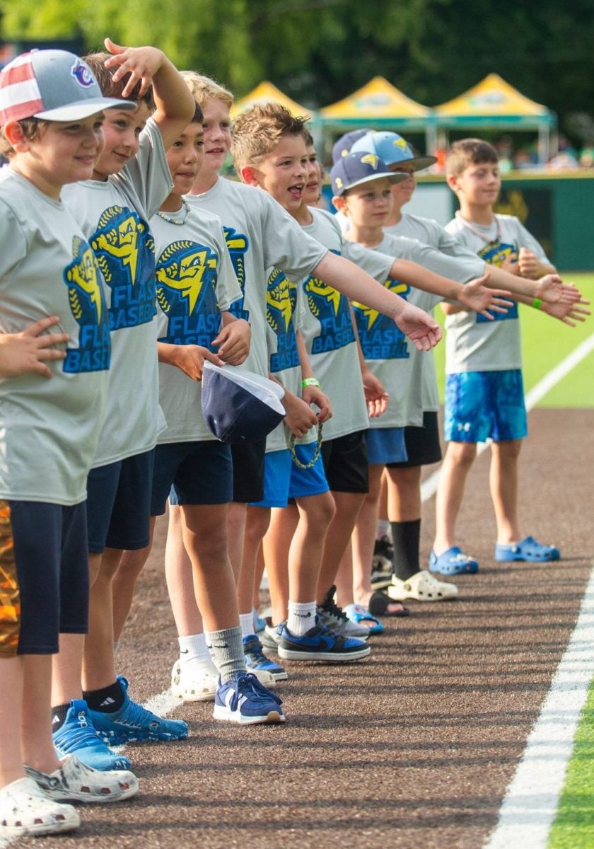 Young baseball players in uniform stand in a line on a baseball field, facing right.